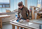 A person wearing safety equipment sands a wooden window frame in a workshop.