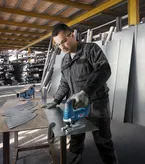 A person wearing safety equipment cuts sheet metal with a cordless jigsaw in a workshop.