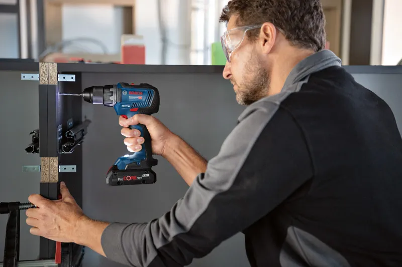 A person wearing safety equipment drills into a cabinet frame with a cordless power drill.