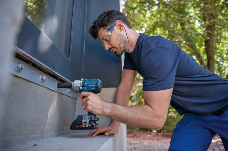 A person wearing safety equipment uses a cordless impact wrench to fasten bolts on a metal frame.