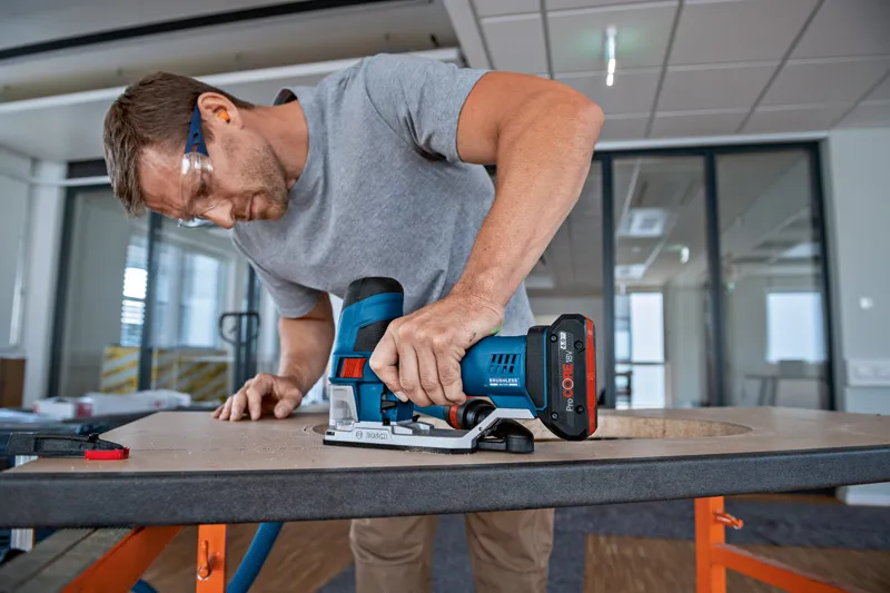 A person wearing safety equipment uses a cordless jigsaw to cut a hole in a wooden board.