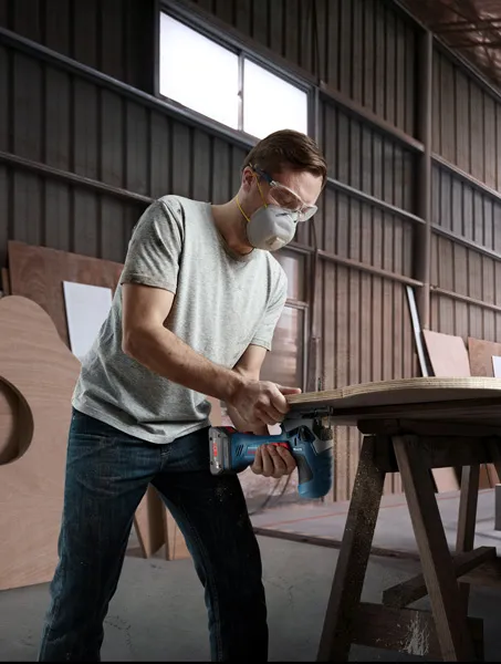 A person wearing safety equipment cuts wood with a cordless jigsaw in a workshop.