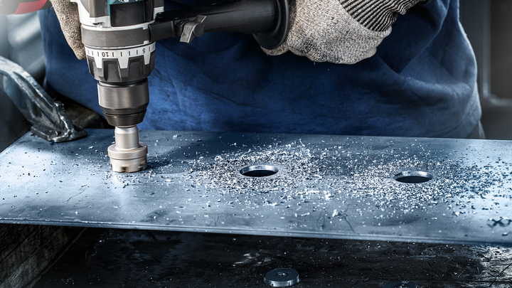 Worker wearing safety equipment drills a large hole in a metal plate.