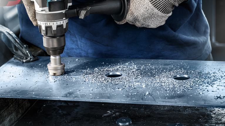 Worker wearing safety equipment drills a large hole in a metal plate.