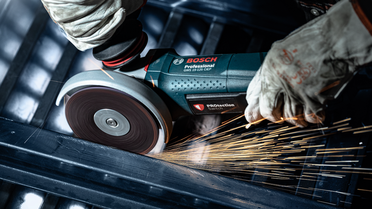 Person wearing safety equipment uses an angle grinder to cut metal, producing sparks.