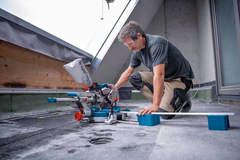 A person wearing safety equipment cuts a board with a cordless mitre saw on a rooftop.