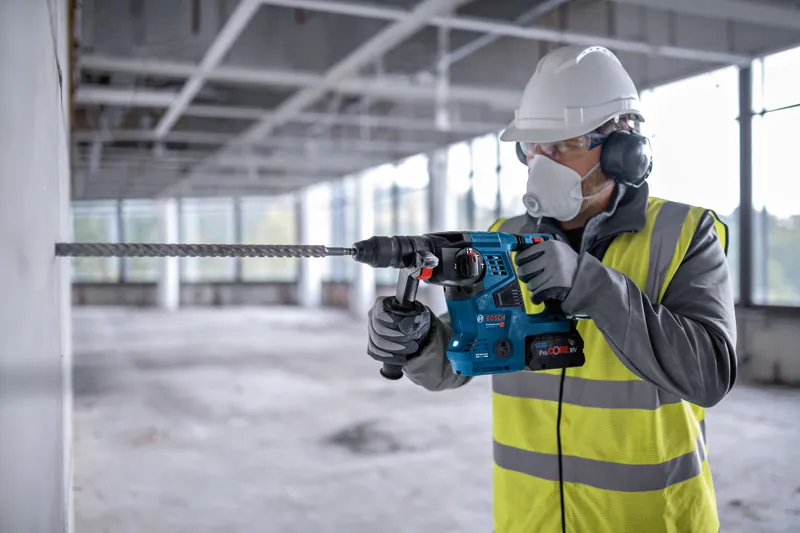 A person wearing safety equipment drills into concrete using a cordless rotary hammer.