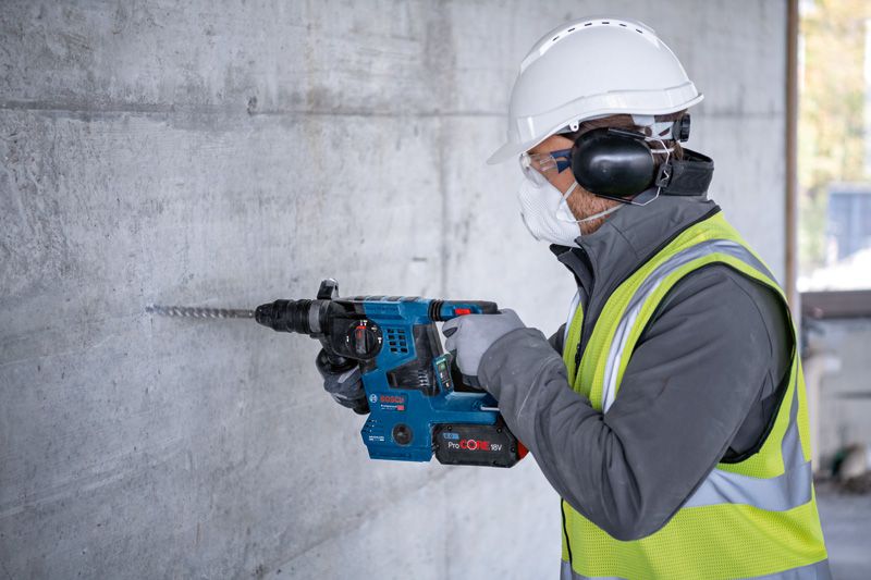 A person wearing safety equipment drills into a concrete wall with a cordless rotary hammer.