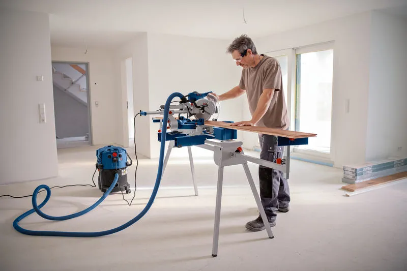 A person wearing safety equipment cuts wood on a mitre saw in a bright, unfinished room.