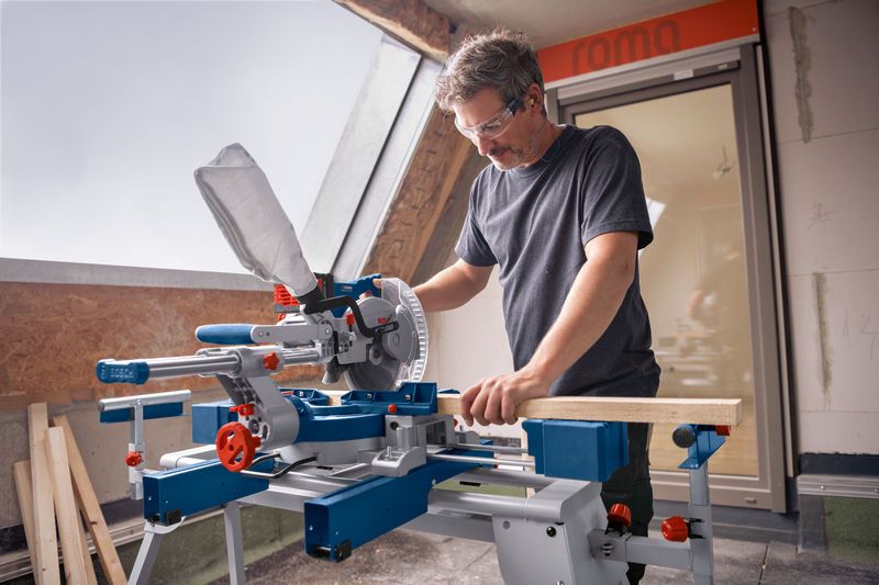 A person wearing safety equipment operates a cordless mitre saw to cut wood indoors.
