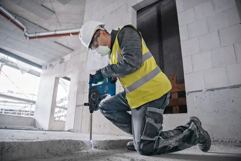 A person wearing safety equipment uses a hammer drill to break concrete on a construction site.
