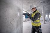 A person wearing safety equipment drills into a concrete wall at a construction site.
