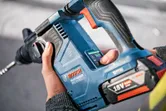 A person wearing safety equipment operates a cordless rotary hammer drill.