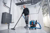 A person wearing safety equipment uses a cordless dust extractor to clean a tiled floor.