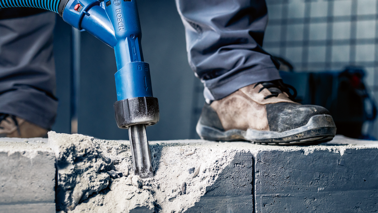 Person wearing safety equipment breaks concrete with a power tool.