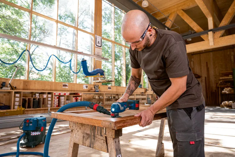 A person wearing safety equipment sands wood on a workbench using a power tool.