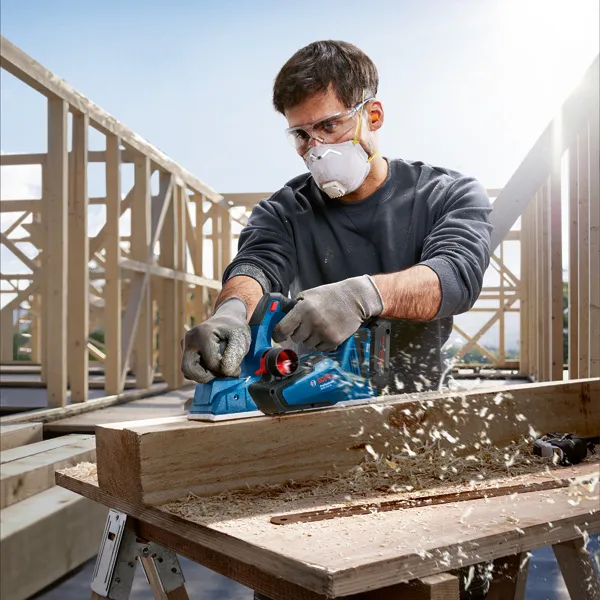 A person wearing safety equipment planes a wooden beam at a construction site.