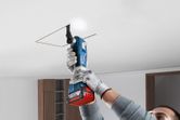 A person wearing safety equipment cuts a square opening in the ceiling with a cordless power tool.