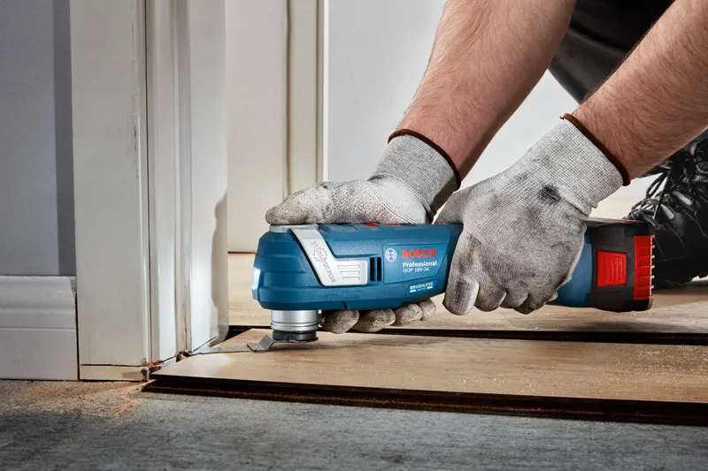 A person wearing safety equipment trims flooring near a door frame with a multitool.