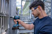 A person wearing safety equipment uses a cordless impact wrench on a metal structure.