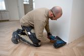 A person wearing safety equipment uses a cordless wood nailer to install floor trim.