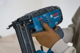 A person wearing safety equipment adjusts a cordless wood nailer.