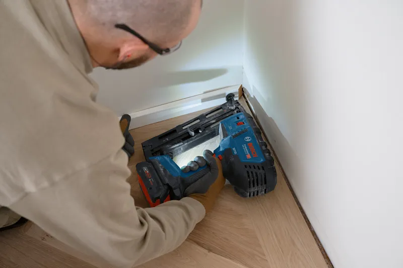 A person wearing safety equipment uses a cordless wood nailer on floor trim.