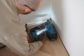A person wearing safety equipment uses a cordless finish nailer on floor trim.