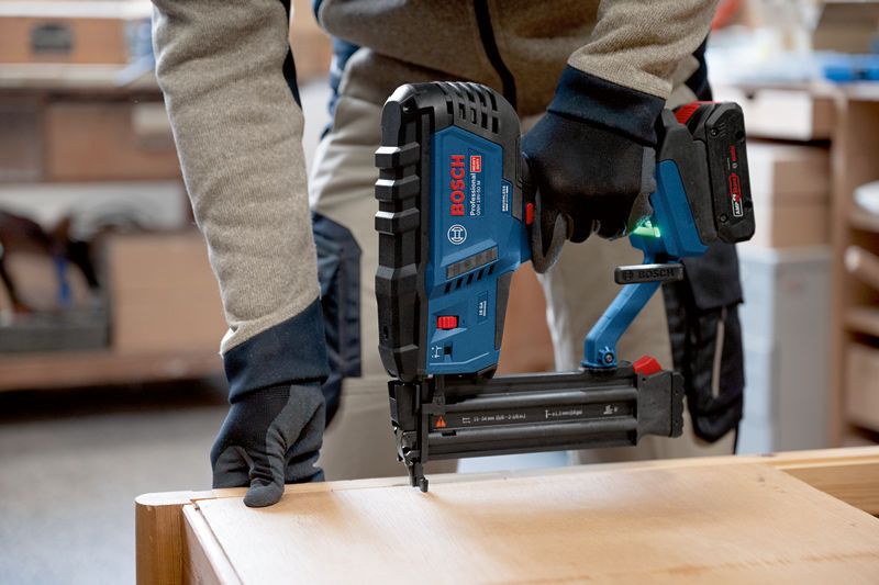 A person wearing safety equipment uses a cordless wood nailer on a wooden surface.