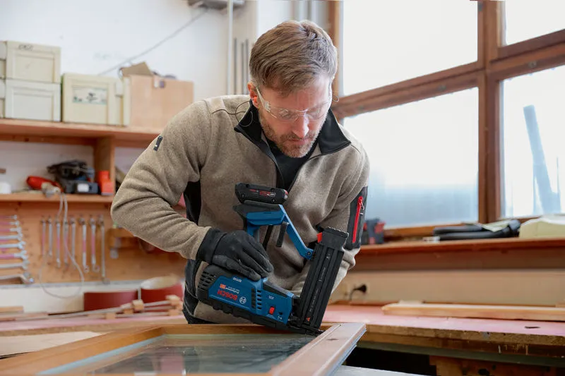 A person wearing safety equipment uses a cordless wood nailer on a window frame.