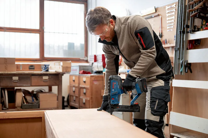 A person wearing safety equipment uses a cordless wood stapler on a wooden board.