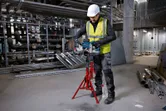 A person wearing safety equipment cuts a metal pipe with a cordless band saw on a tripod.