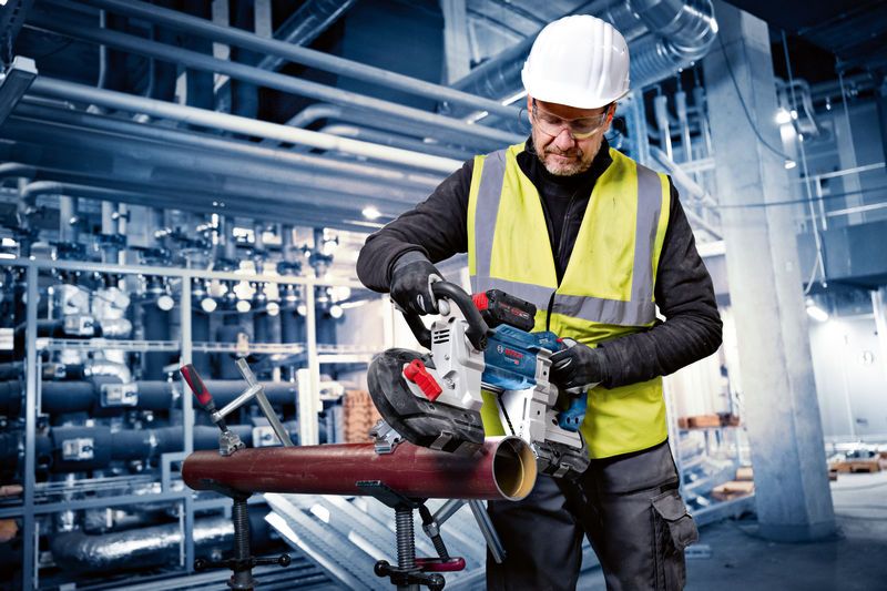 A person wearing safety equipment uses a cordless band saw to cut a metal pipe.