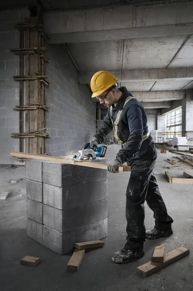 A person wearing safety equipment cuts wood with a circular saw in a construction site.
