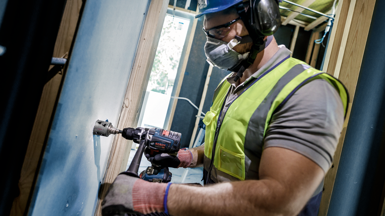 Person wearing safety equipment drills into a wall stud inside a building under construction.