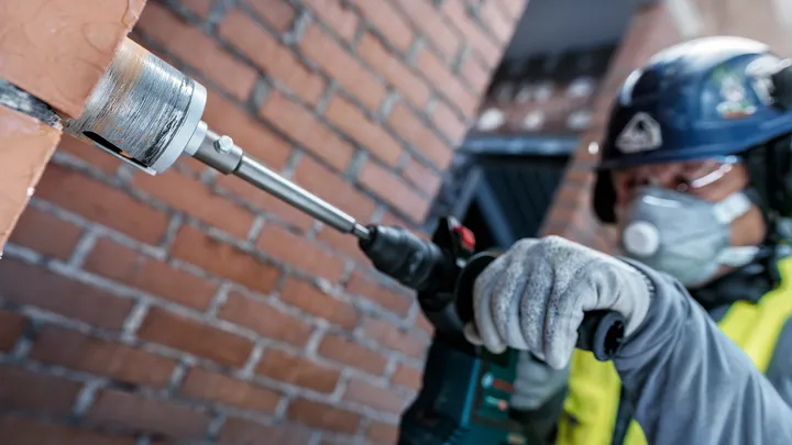 Person wearing safety equipment drills a hole into a brick wall.