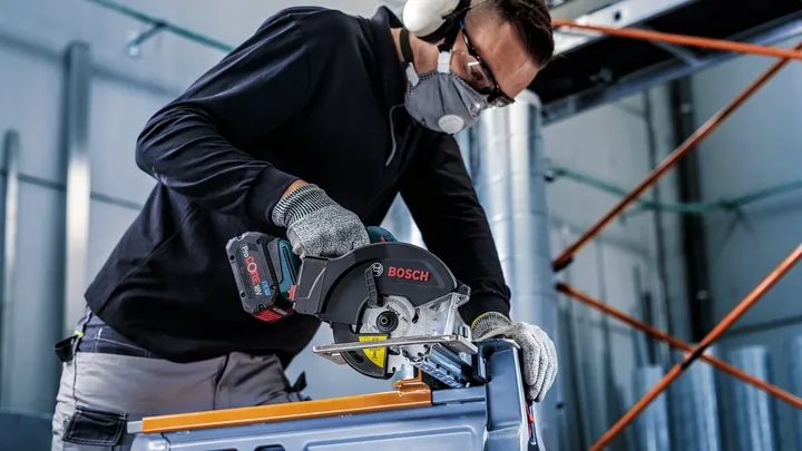 A person wearing safety equipment uses a circular saw to cut metal.