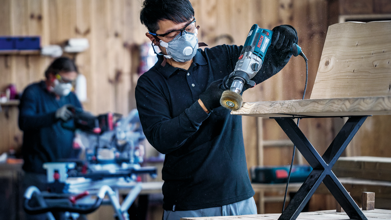 A person wearing safety equipment polishes a wooden chair in a workshop.