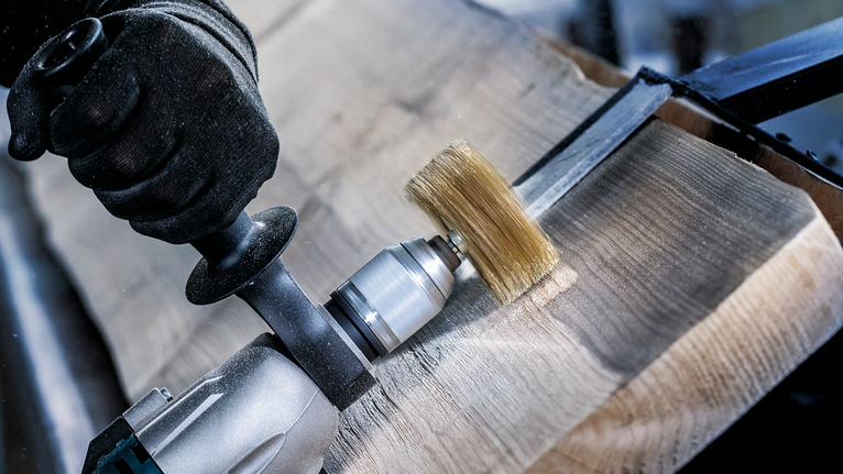 Person wearing safety equipment uses a rotary tool with a brush to treat a wooden surface.