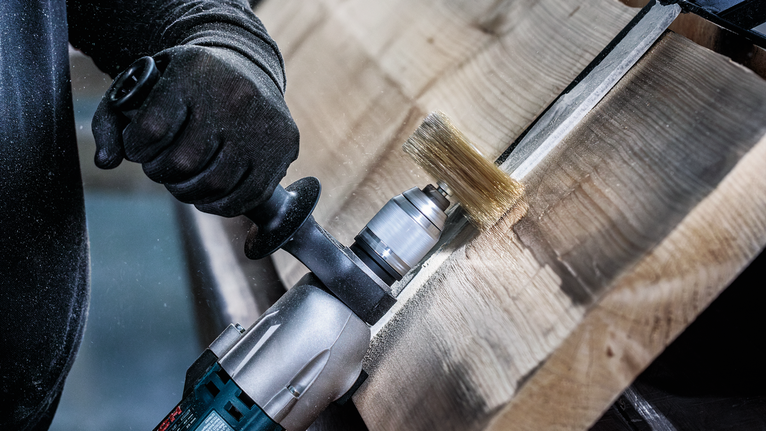 Person wearing safety equipment uses a power tool with a wire brush to clean wood.