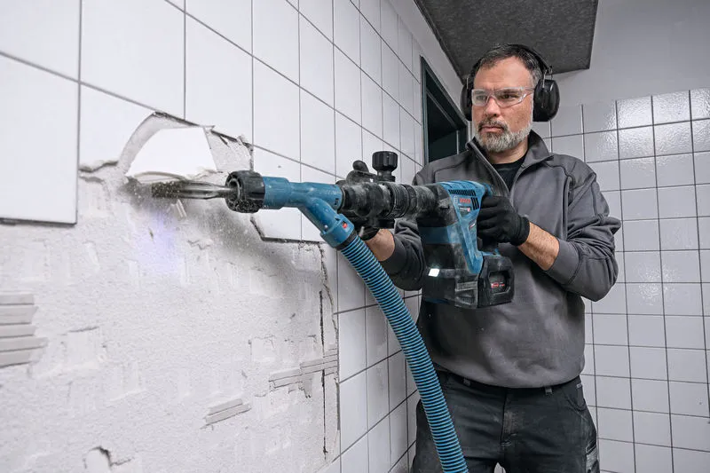 A person wearing safety equipment uses a cordless demolition hammer to break wall tiles.