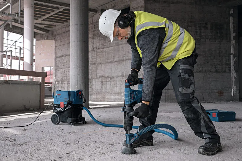 A person wearing safety equipment drills concrete with a dust extraction attachment.