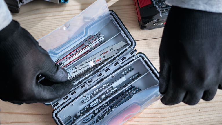 Person wearing safety equipment selects saw blades from a plastic case on a wooden surface.