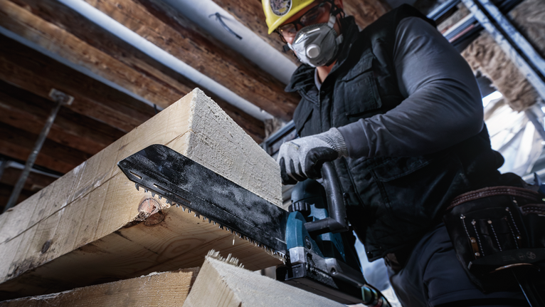 Person wearing safety equipment cuts thick wooden beams using a reciprocating saw.