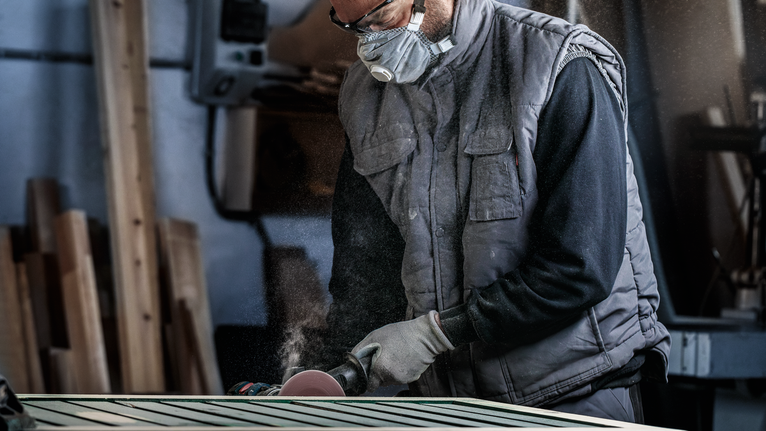 A person wearing safety equipment sands a wooden surface with a power tool.