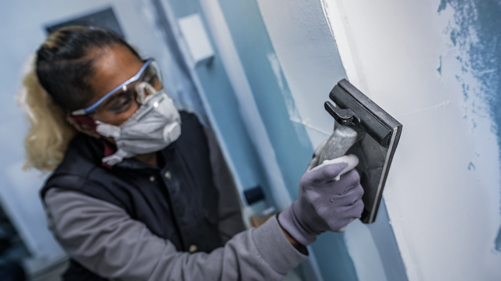 Person wearing safety equipment smooths a wall surface with a sanding tool.