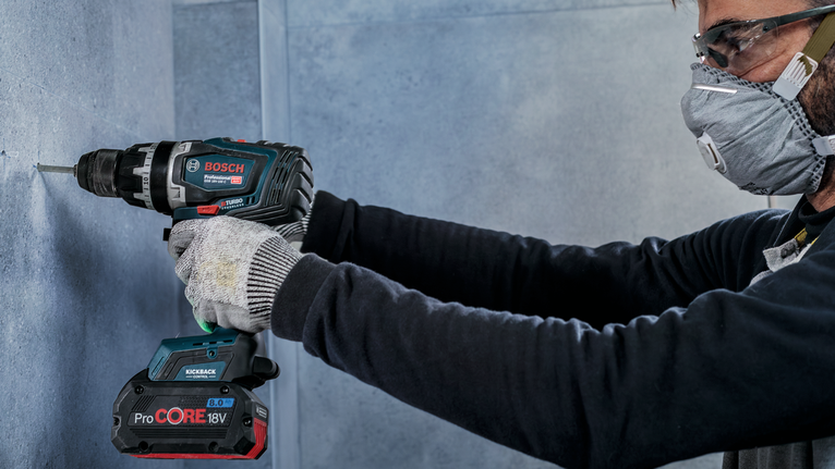 A person wearing safety equipment drills into a concrete wall.