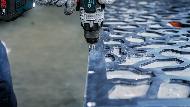 Person wearing safety equipment drills into a patterned metal sheet.