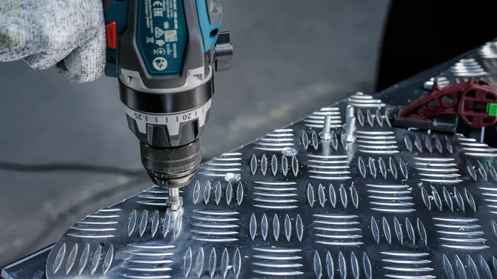 Person wearing safety equipment uses a power drill to fasten screws into a metal plate.