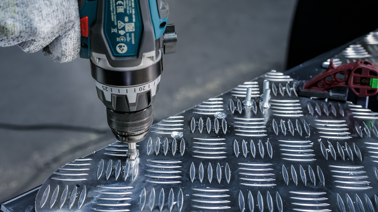 Person wearing safety equipment uses a power drill to fasten screws into a metal plate.